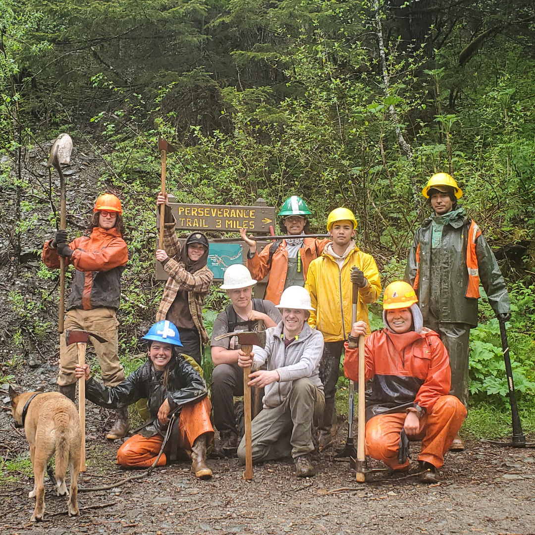 A group of 9 trail crew members wearing hard hats, rain jackets, and rain pants 